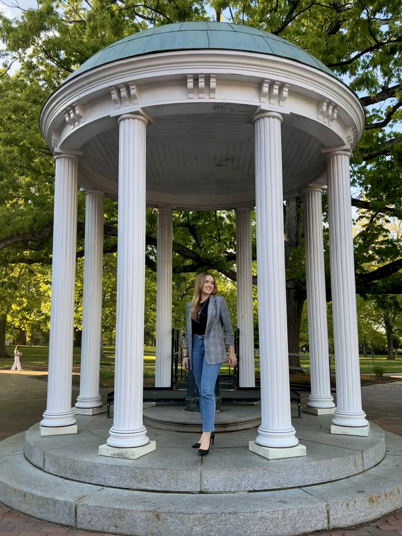 Picture of young women in light jeans, black tucked in shirt and a checkered gray blazer. She is leaning on her back leg with her face toward the side. She is in a gazebo with columns around her out in a park setting. 