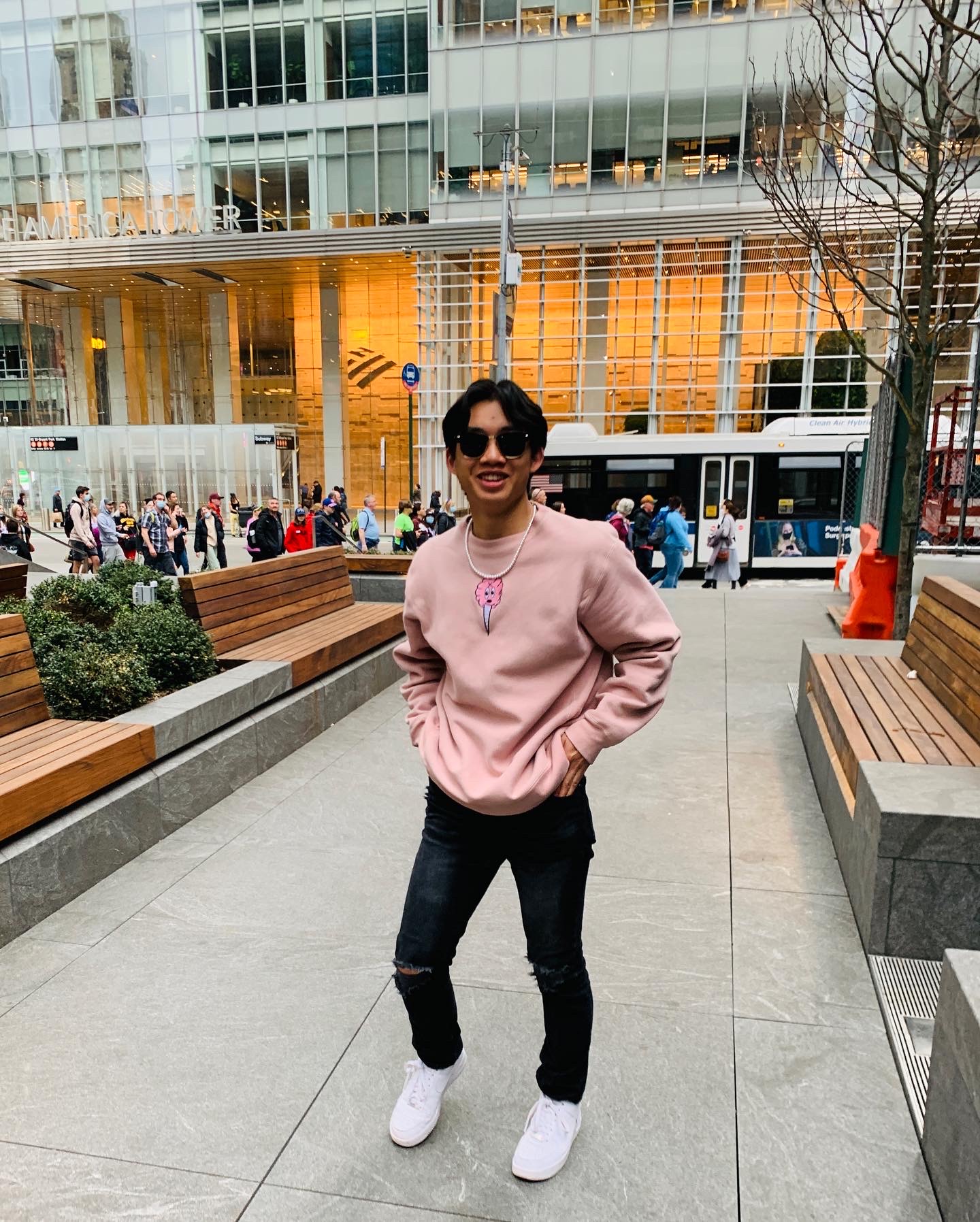 Picture of young male student with a pink sweatshirt, dark jeans, white sneakers and sunglasses on. Student is in the walkway between several benches in a city.