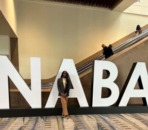 Picture of a young women student standing in front of large letters spelling out NABA. She is wearing a gray blazer with a tucked in beige shirt and fawn colored pants with heels. She is standing facing the photographer with her hands crossed in front.